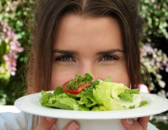 Young woman looking to camera holding a plate of salad in front of her face.